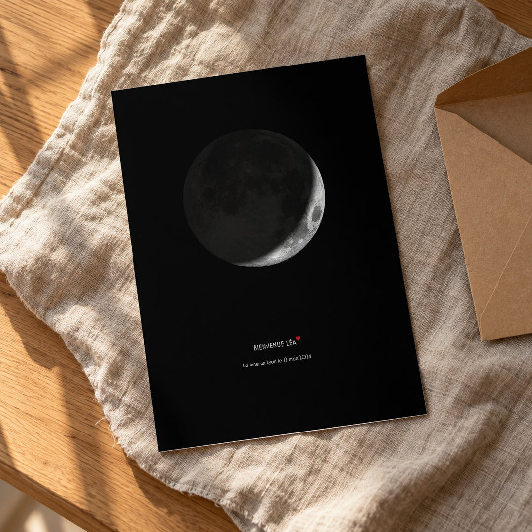 Affiche Lune « Bienvenue Léa » imprimée, posée à plat sur un plaid en lin à côté de son enveloppe rigide